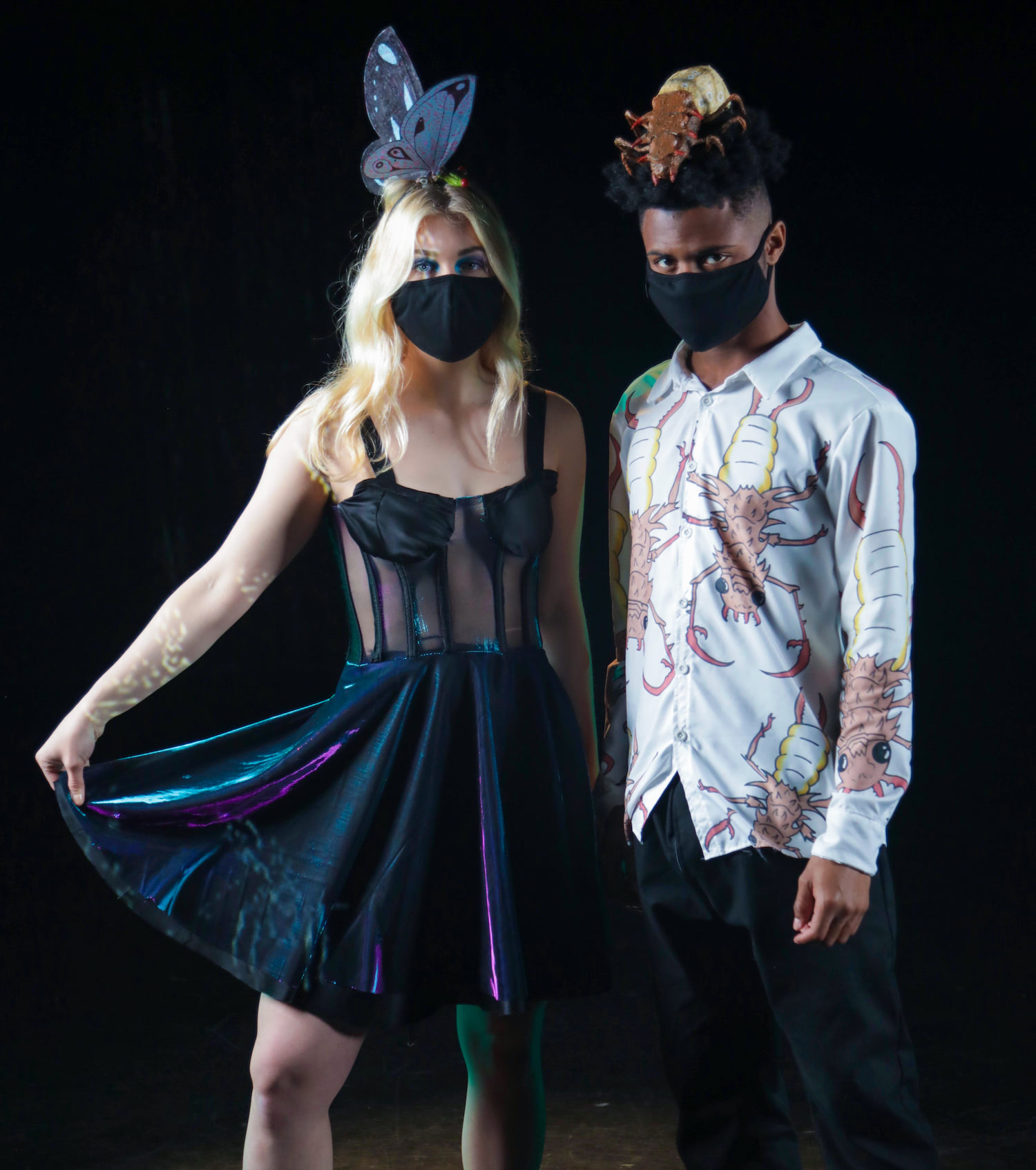 The image depicts the two models next to each other in the full outfits created by the artist. The woman wears a black dress, with purple/blue iridescence, and a crown of a butterfly being eaten. The man has a button up and slacks. The slacks are black with green/blue iridescent pockets. The shirt is white with a large all-over pattern of brown and yellow insects with large claws. He wears a sculpture of one of these insects on his head like a hat.