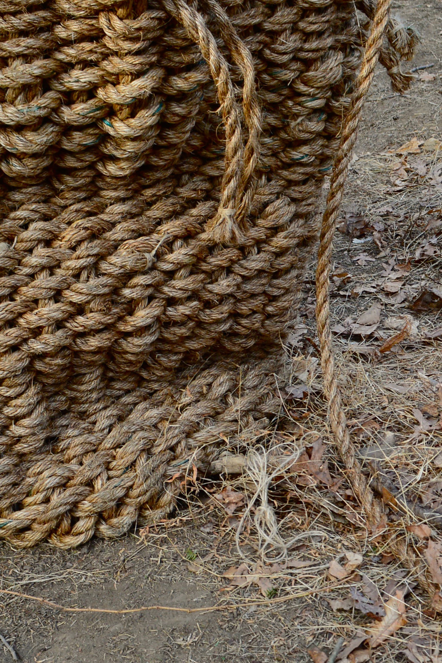 The base, which folds in on itself several times due to the weight of the basket, is situated on the left hand side of the photograph. Some of the hanging rope used as weavers, or horizontal weaving material, is tied with jute twine to ensure the edges do not fray. The sculpture is grounding itself, pulling the surrounding dead grasses and leaves toward its base. A harmonious relationship exists between the muted yellow and brown tones of the basket and the surrounding environment.
