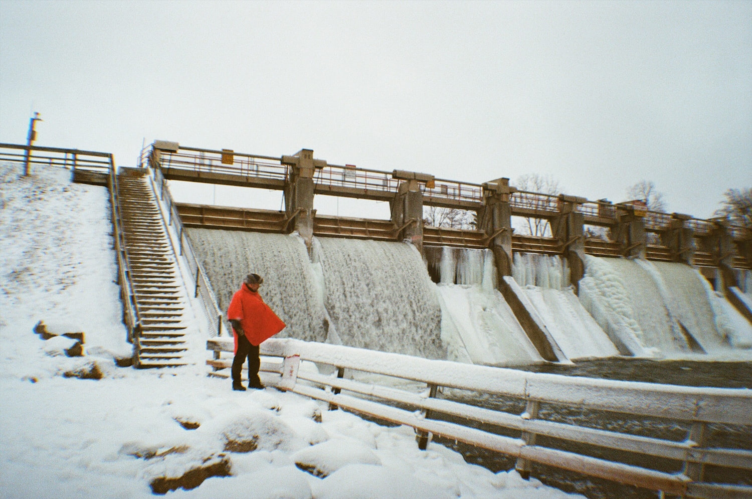 The half-frozen Barton Dam roars behind a man in an orange poncho, blowing from the strong winter wind.