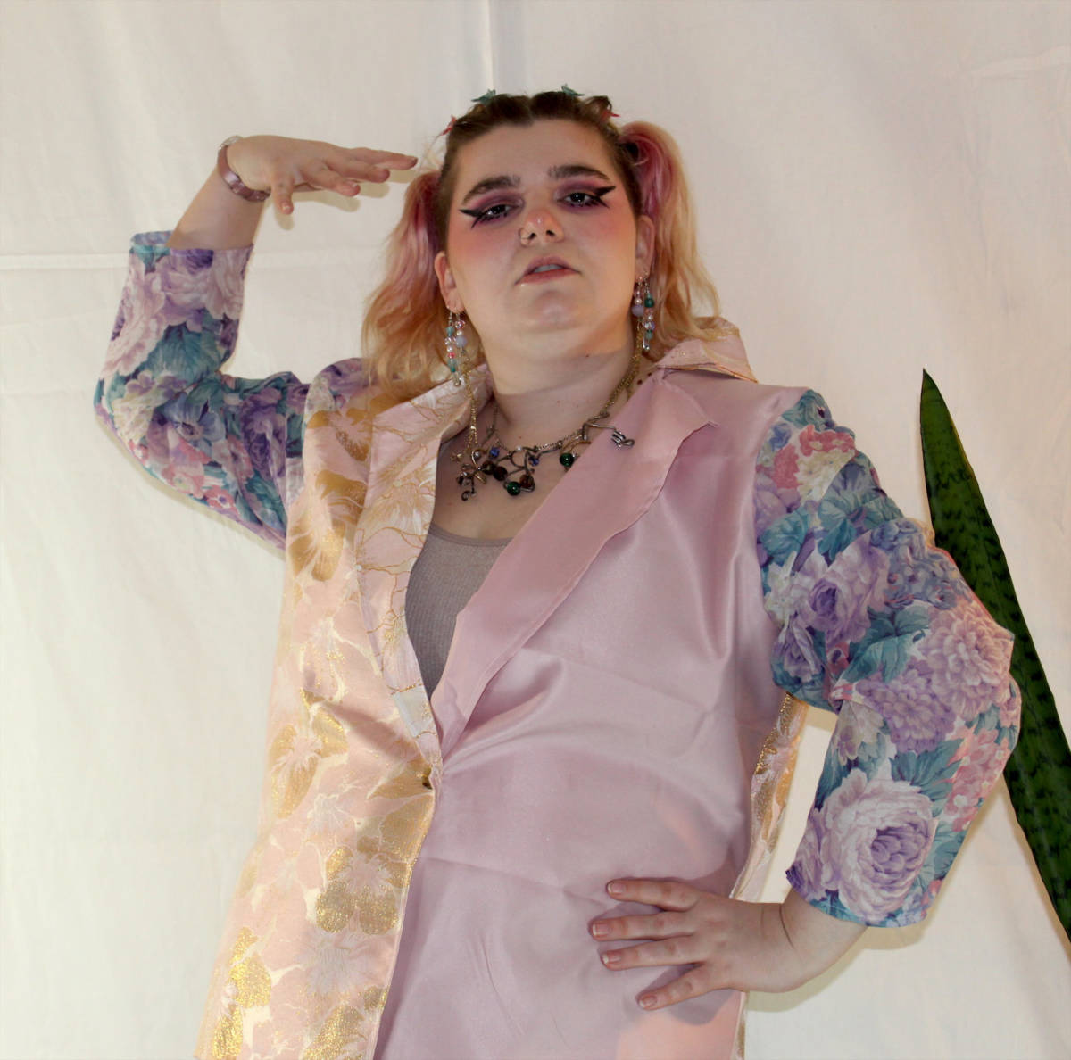 The artist is wearing wearable two in a closeup. The piece is a suit jacket which features a pink satin fabric with a gold metallic floral design as the body of the work. The sleeves are a floral fabric with shades of green, purple, and pink.