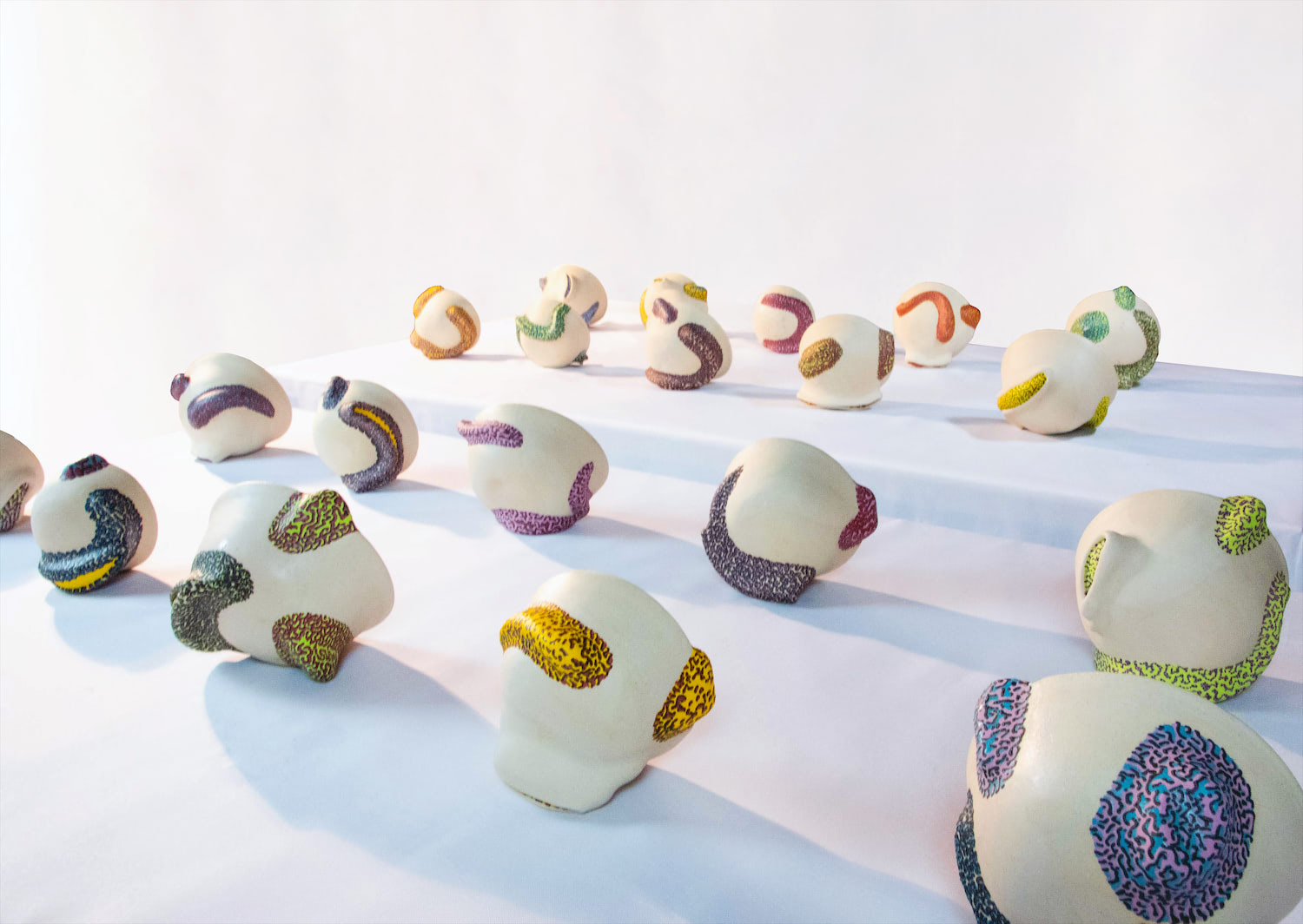 An image of the exteriors of many white bowl forms with colorful sculptural additions displayed on 2 rectangular tables with white table cloths.