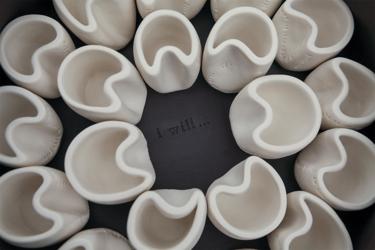 High angle image of twenty white porcelain cups in black porcelain carrier.  The cups are centered around the statement “I will…” that is stamped onto the center of the carrier.