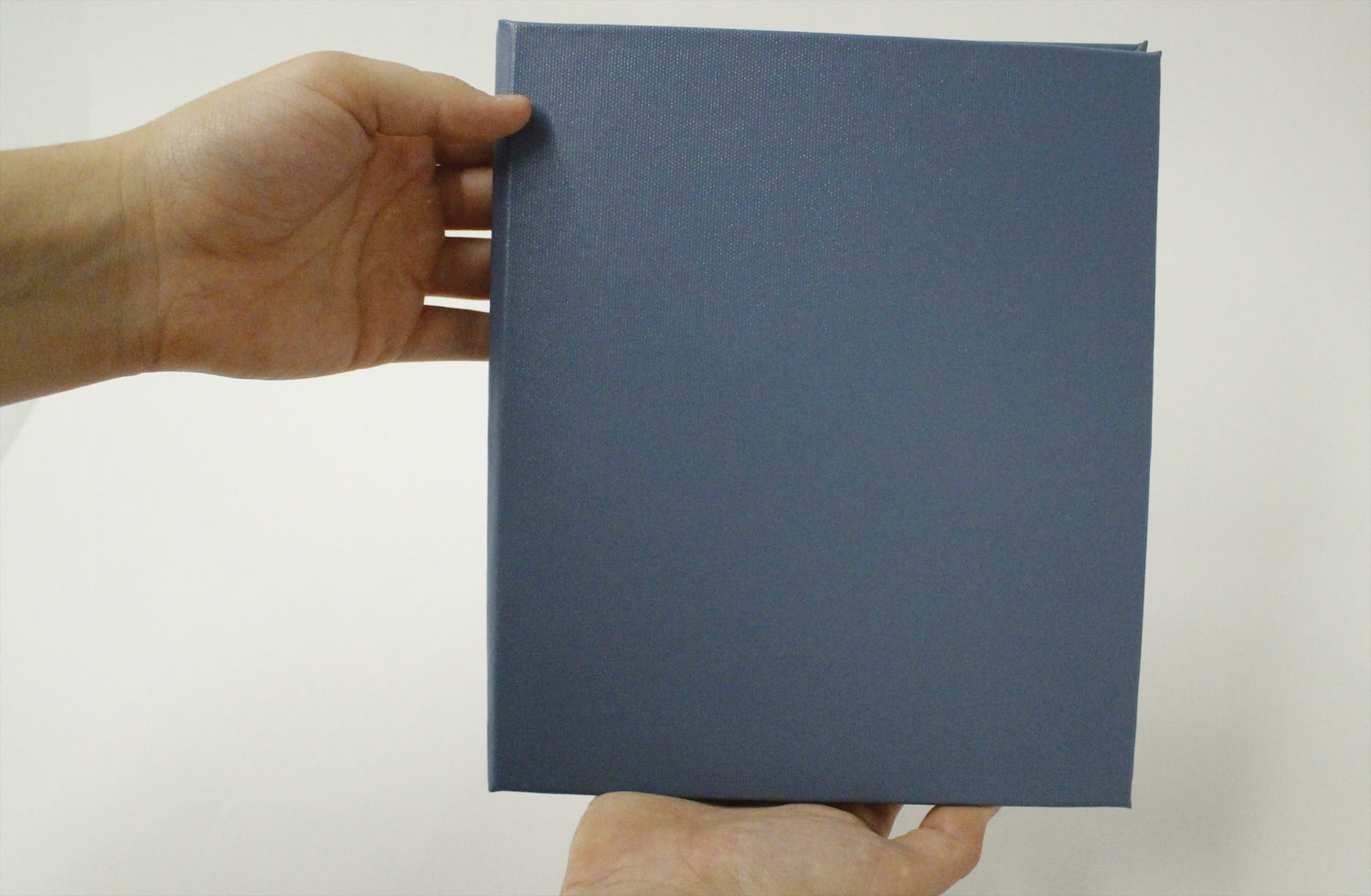 The artist's hands hold a closed self-bound book, showing its blue cover.