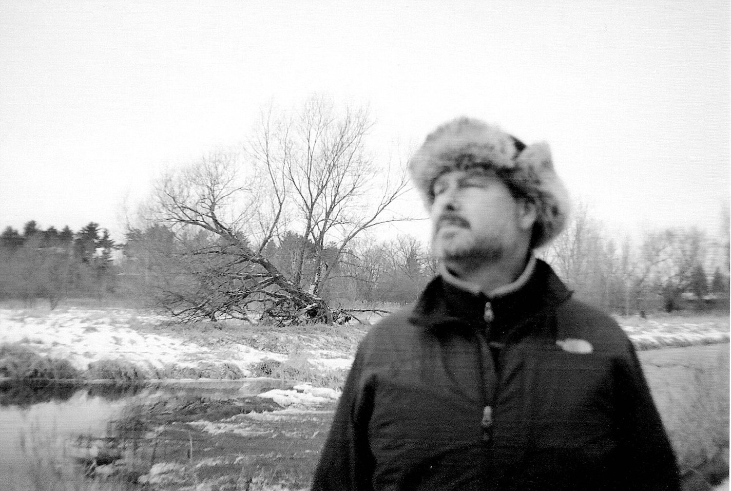A man in a furry winter hat gazes at the sky with a bare tree and partially frozen river behind him.