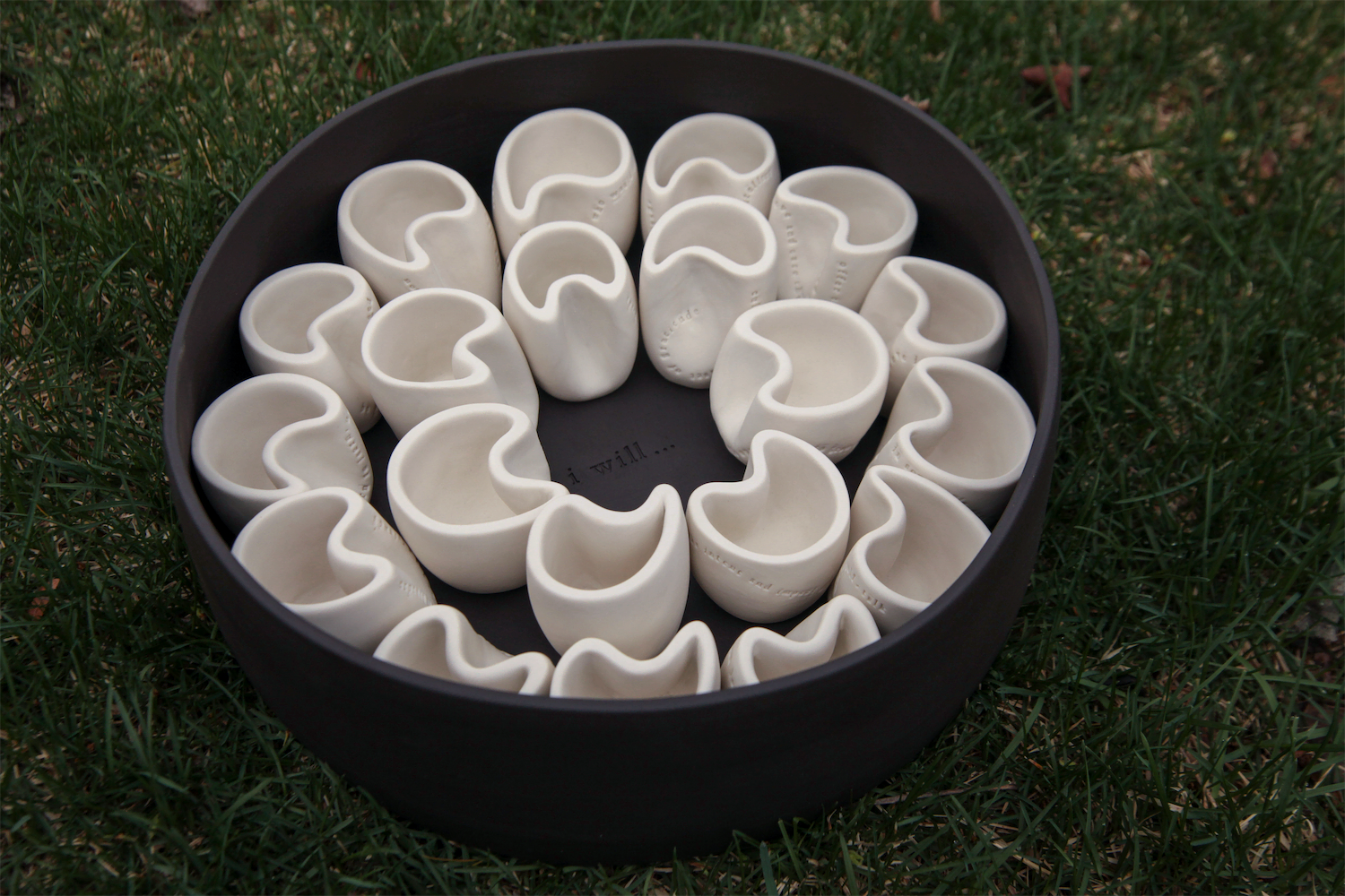 Twenty white porcelain cups in black porcelain carrier.  The cups are centered around the phrase “I will…” that is stamped onto the center of the carrier.