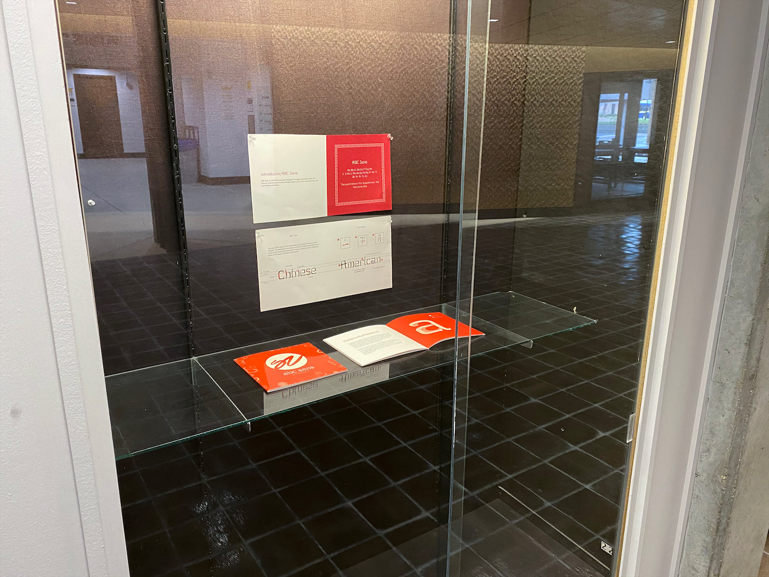 Two guide books, one closed and one open, sit on a glass shelf in a display case it. Above them hang two spreads from the inside of the booklet.