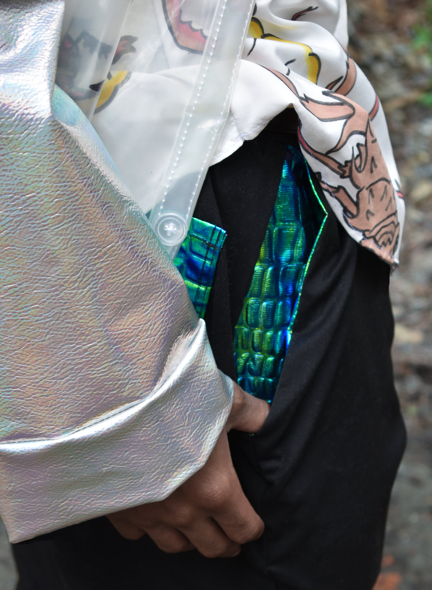 A close up of the man's jacket sleeve and pants pocket. The inside of the pocket is a scaly green/blue material that shines in the light. The cuff of the jacket sleeve is a silvery iridescent material.
