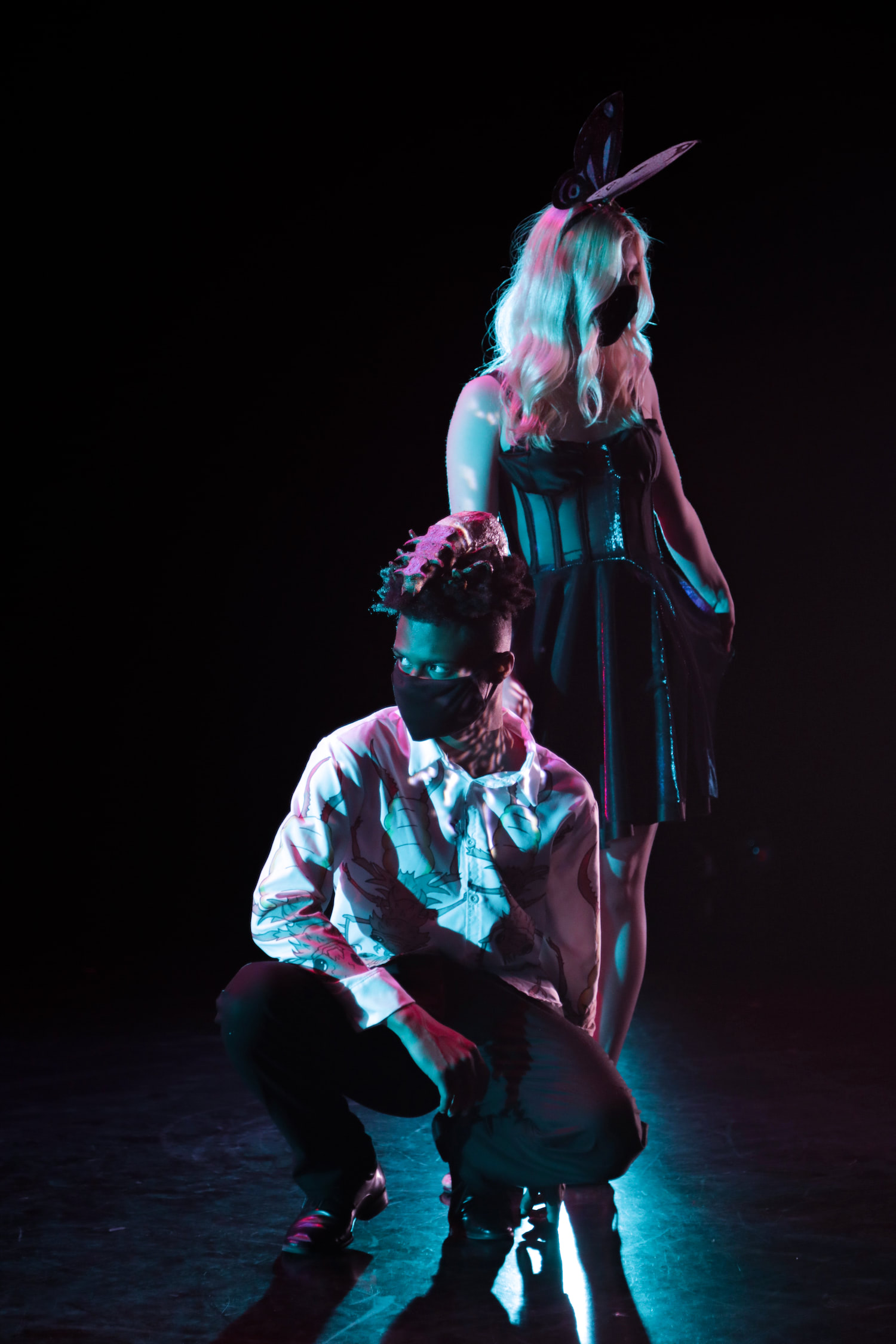 This image is of the models in special studio lighting. The woman stands next to the crouching man. They are bathed in purple and blue light.
