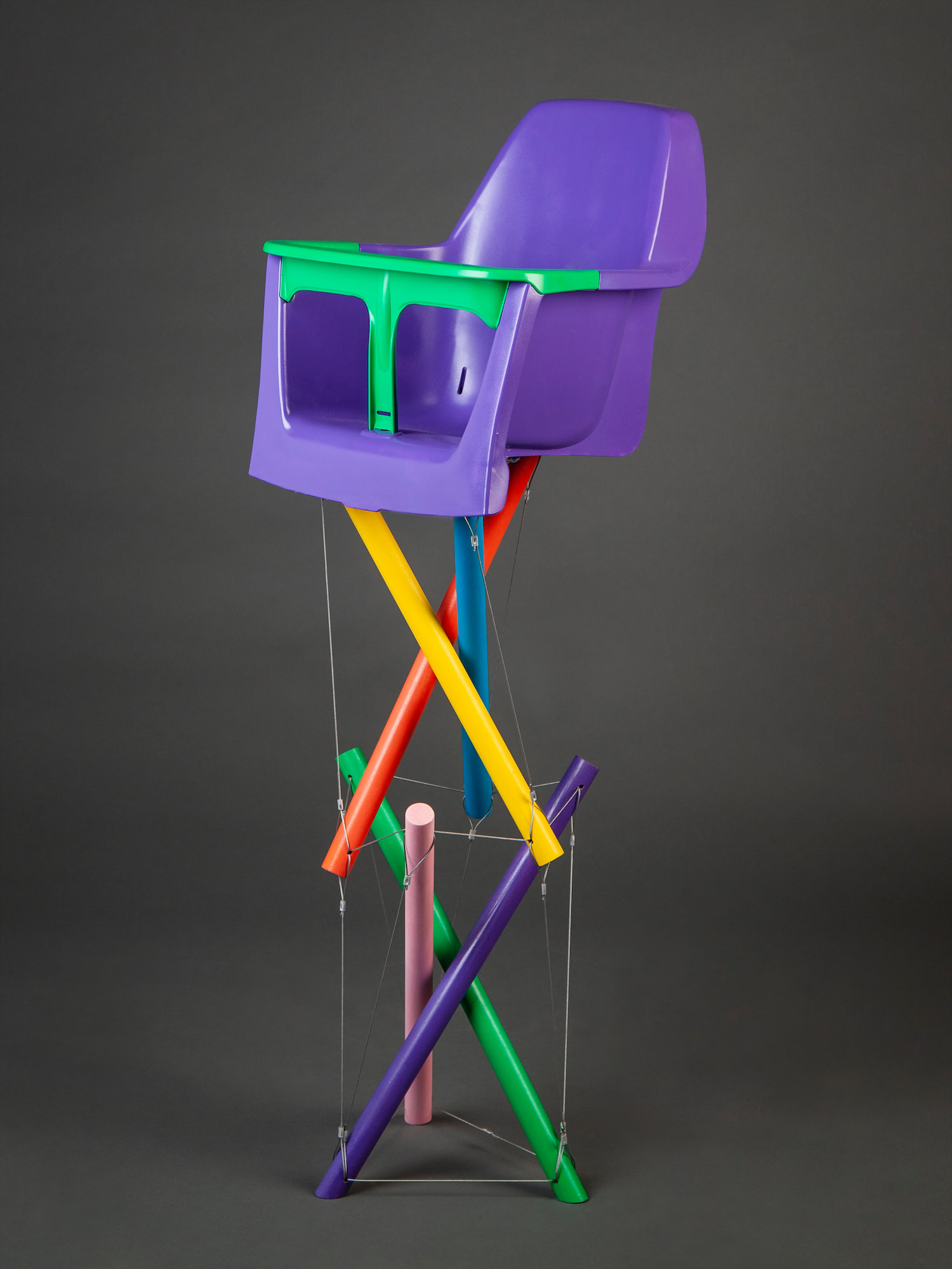 Tension Highchair. A green and purple painted plastic chair on top of six painted round legs, each suspended by cable and tilted at 30 degrees.