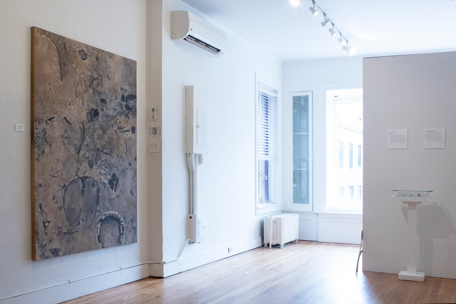 A gallery space with white walls and wood floors, exhibiting the single large painting next to the window of the gallery behind the rock display and artist statement.