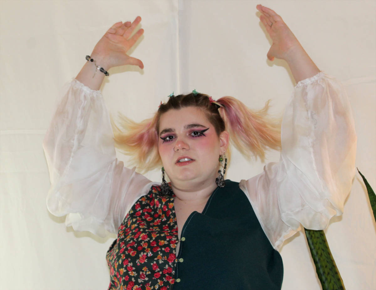 The artist is featured in a closeup where they are wearing wearable one. The artist's arms are raised showing off the flow of the see-through sleeves of the top. The right side of the top is a deep green, while the left side is a pink floral pattern on a similar green background.