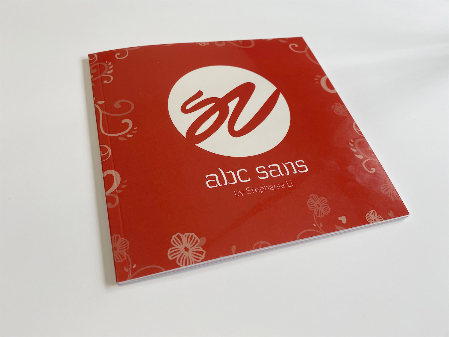 A photo of the cover of the ABC Sans style guide booklet, is photographed at an angle. It has a glossy, red cover, with some muted pink floral designs on its edges.