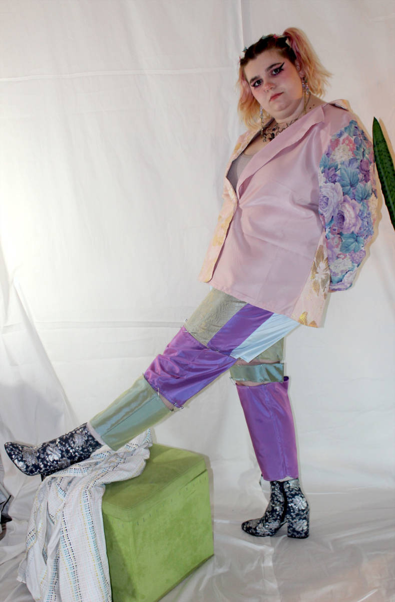 The artist is wearing wearable two in a full body shot. Their left leg is resting straight out on a green cube and both arms are behind the artist. The top is a suit jacket with a pink satin body and purple, pink, and green floral sleeves. The top is paired with pants that are sectioned into separate pieces. The fabrics used in the sections include a metallic purple, gold, and blue which are safety pinned together.