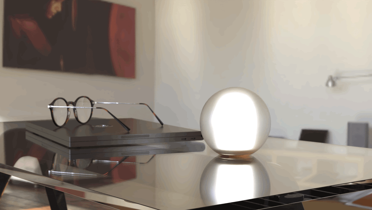 This image shows the sphere-like light turning on and off on a table. The top of the sphere is frosted glass with the bottom part being gold. There is also a laptop and a set of glasses on the glass table.