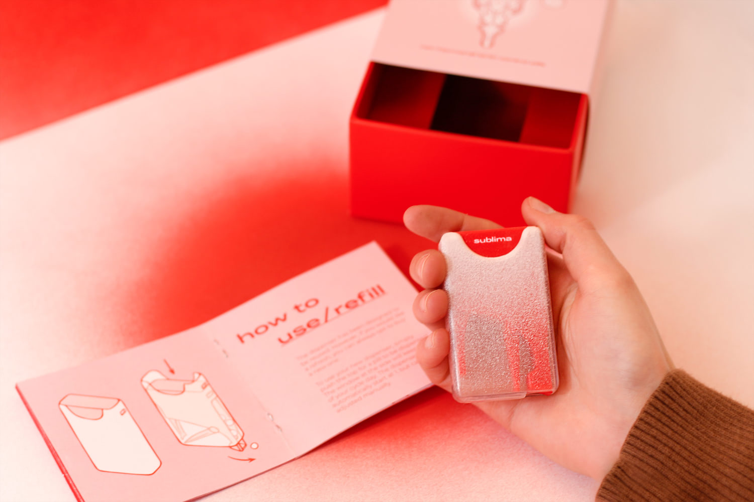 A photo showing the pill dispenser out of the packaging box and in the user's hand. The pill dispenser's exterior body is made out of a semi-transparent material featuring a gradient from light pink to white/transparent. The red interior body can be somewhat seen through the exterior body. The top button of the dispenser, where the user would push for a pill, has the company name 
