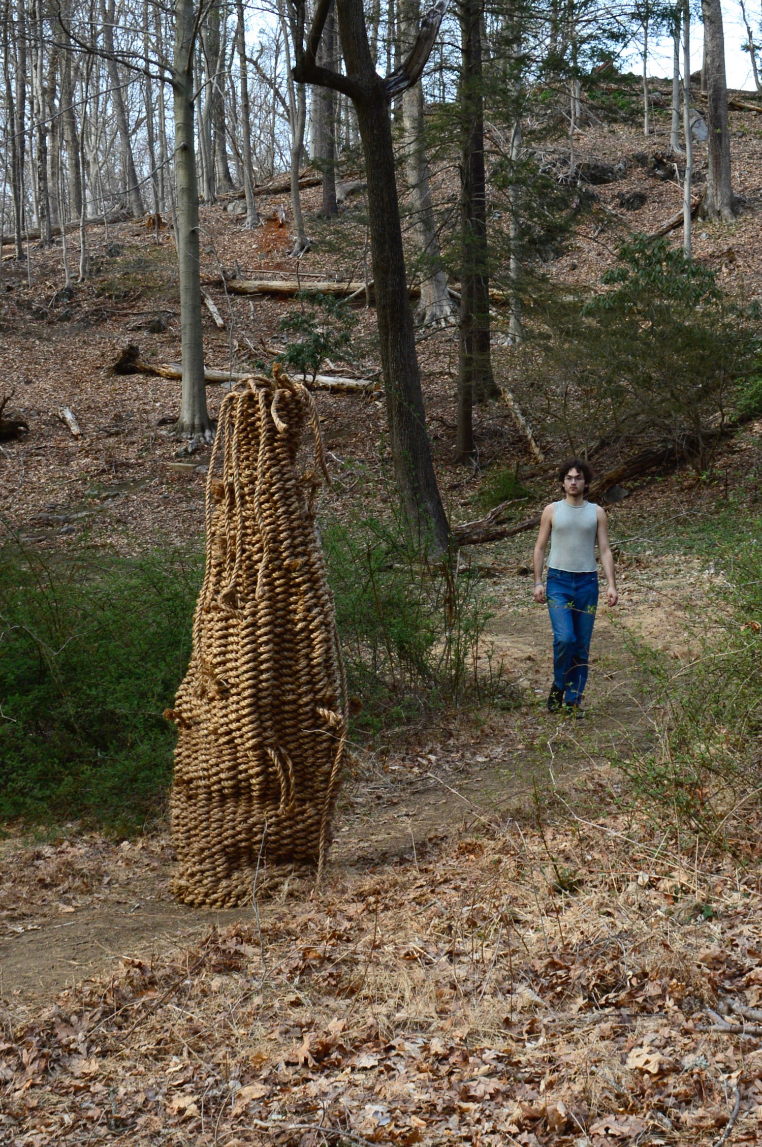 An anthropomorphic basket, or woven column, protrudes from a curving path whose scale is made visible by the artist, who walks toward the viewer from behind the sculpture. Caspar is positioned several feet behind the bushes that line the path and is seen wearing a pale knit tank as well as royal blue jeans. The camera is situated in front of the informational plaque, so the trail marker is not in sight. The left side of the work glows in the sun, so there are deep shadows on its right side.