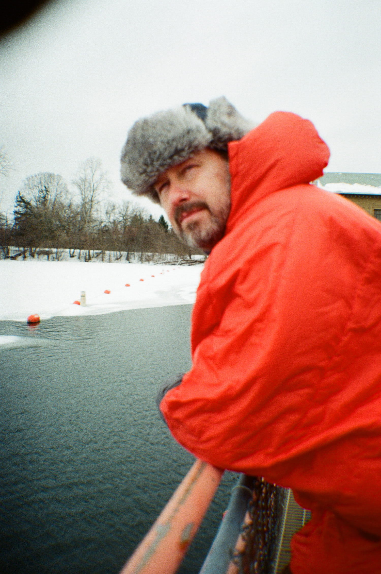 A man in an orange poncho and furry winter hat hunches over the railing atop of Barton Dam, looking out at the frozen lake.