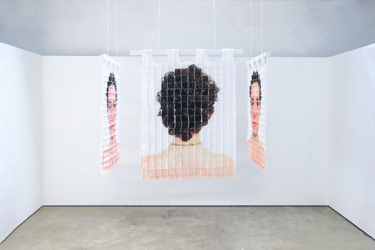 Four quilts hanging in a stark white room, forming a cell-like space. An image of the back of the artist's head, distorted by a grid, faces outward, and is perpendicularly flanked on by a quilt on either side featuring the artist's face distorted by the grid.
