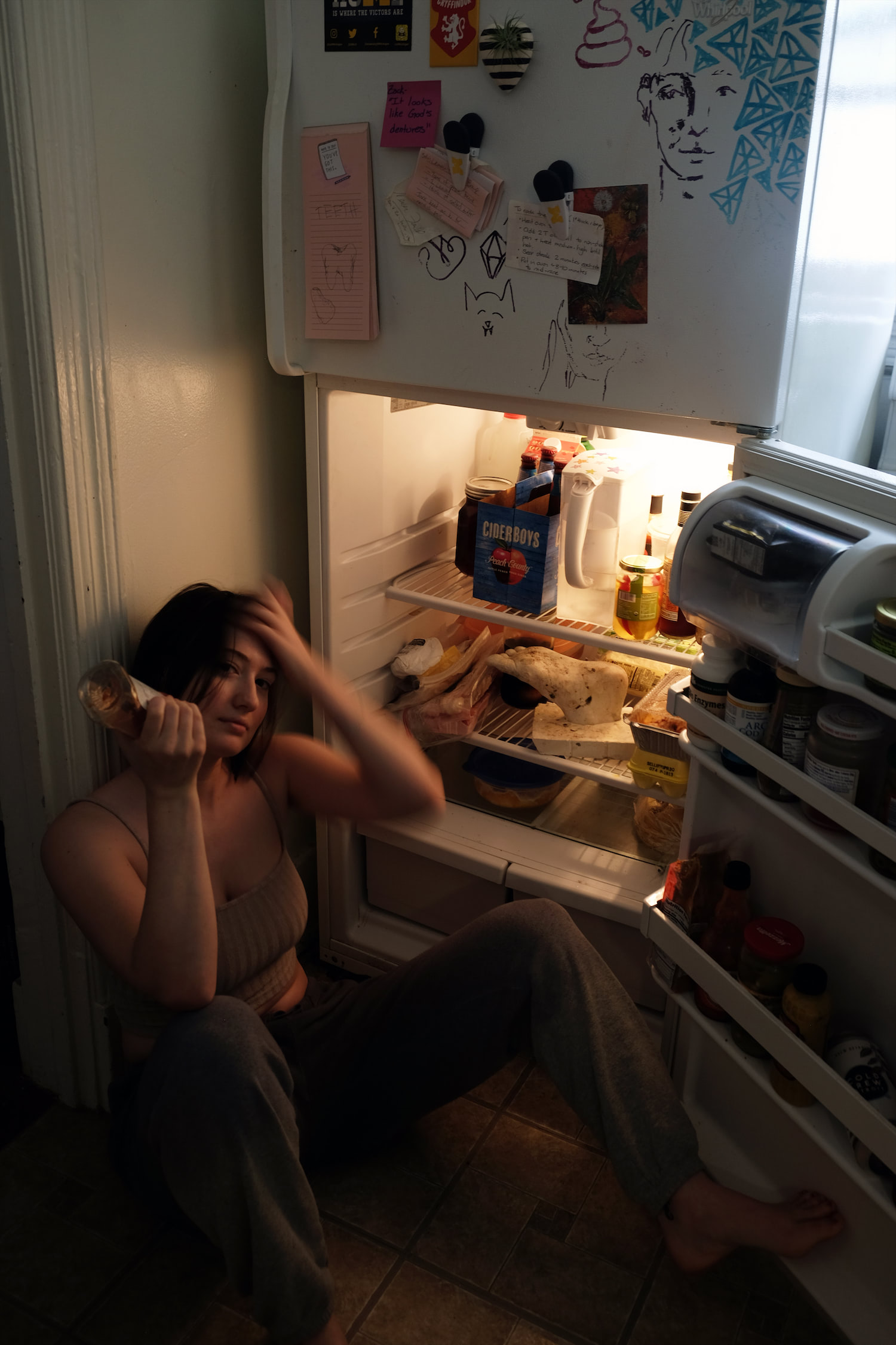 A woman eating ketchup in front of a fridge, in front of a plaster foot covered in bacteria sitting on a fridge shelf.