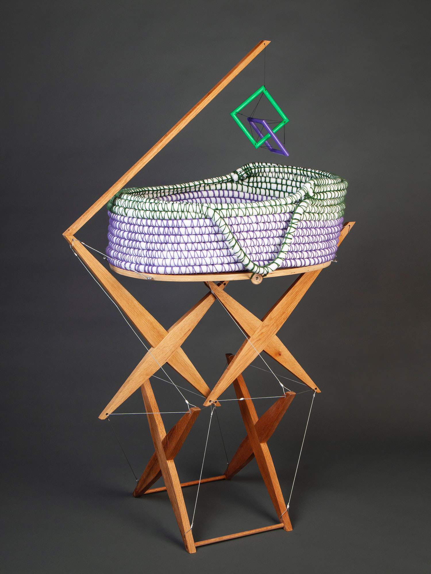 Tensegrity bassinet on a wood structure with a hanging tensegrity mobile and hand crocheted basket. wooden structure is made of two x-like pieces on the ground and two more x-like pieces suspended perpendicularly above them. Steel chords are attached at all four ends of all four x's. The hand crocheted basket is made from coiled 3/4